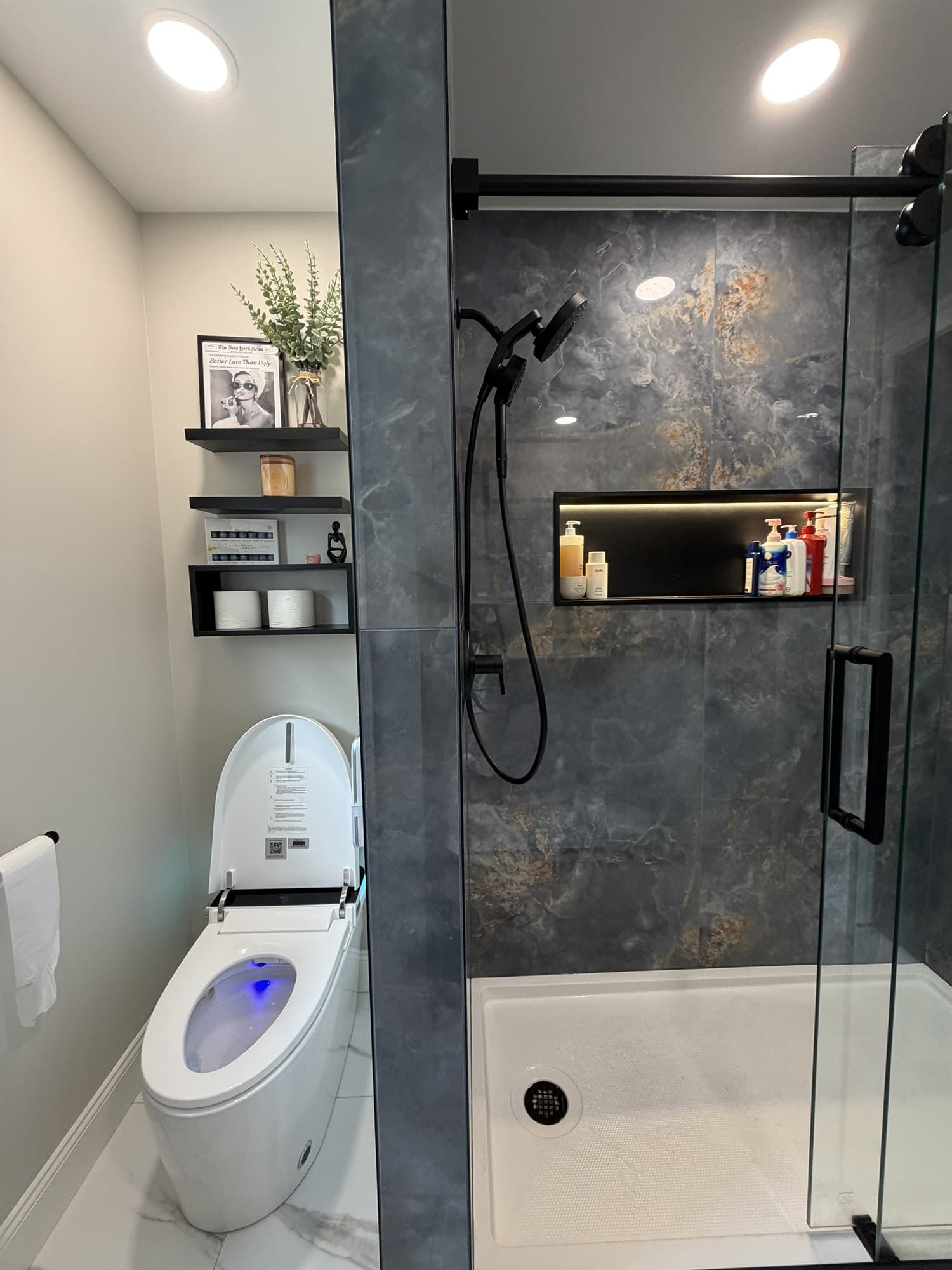 Slate-tiled shower beside the smart toilet and floating black shelves, with marble-look floor tile